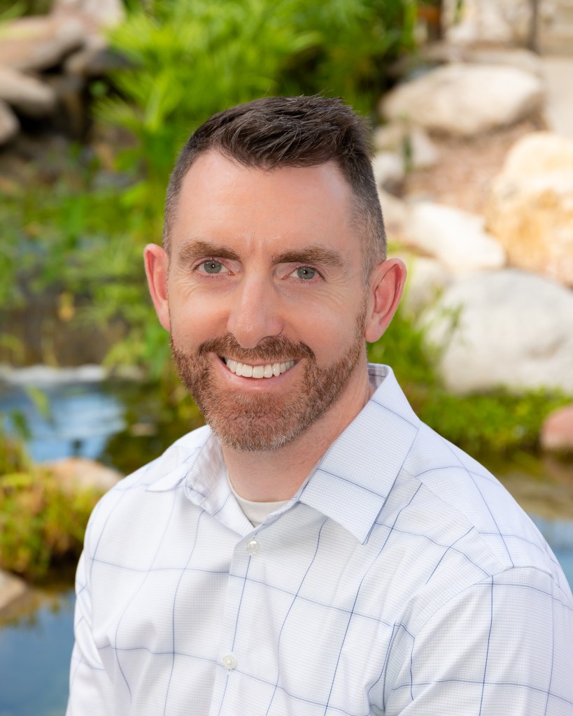 Headshot of Evan Fahy, CPIC. Smiling male with short auburn hair and short beard. Wearing white collared shirt. Natural backdrop of rocks, water, and greenery.
